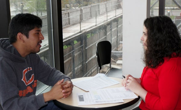 A male student and female advisor sit across from each other at a round table talking. Papers are spread on the table between them.