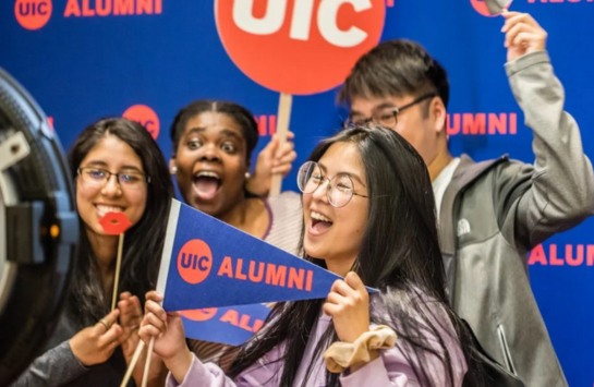 Three women and one man hold small UIC alumni banners while posing in front of a blue background with the words UIC Alumni printed on it in red. They are celebrating while taking a group photo.